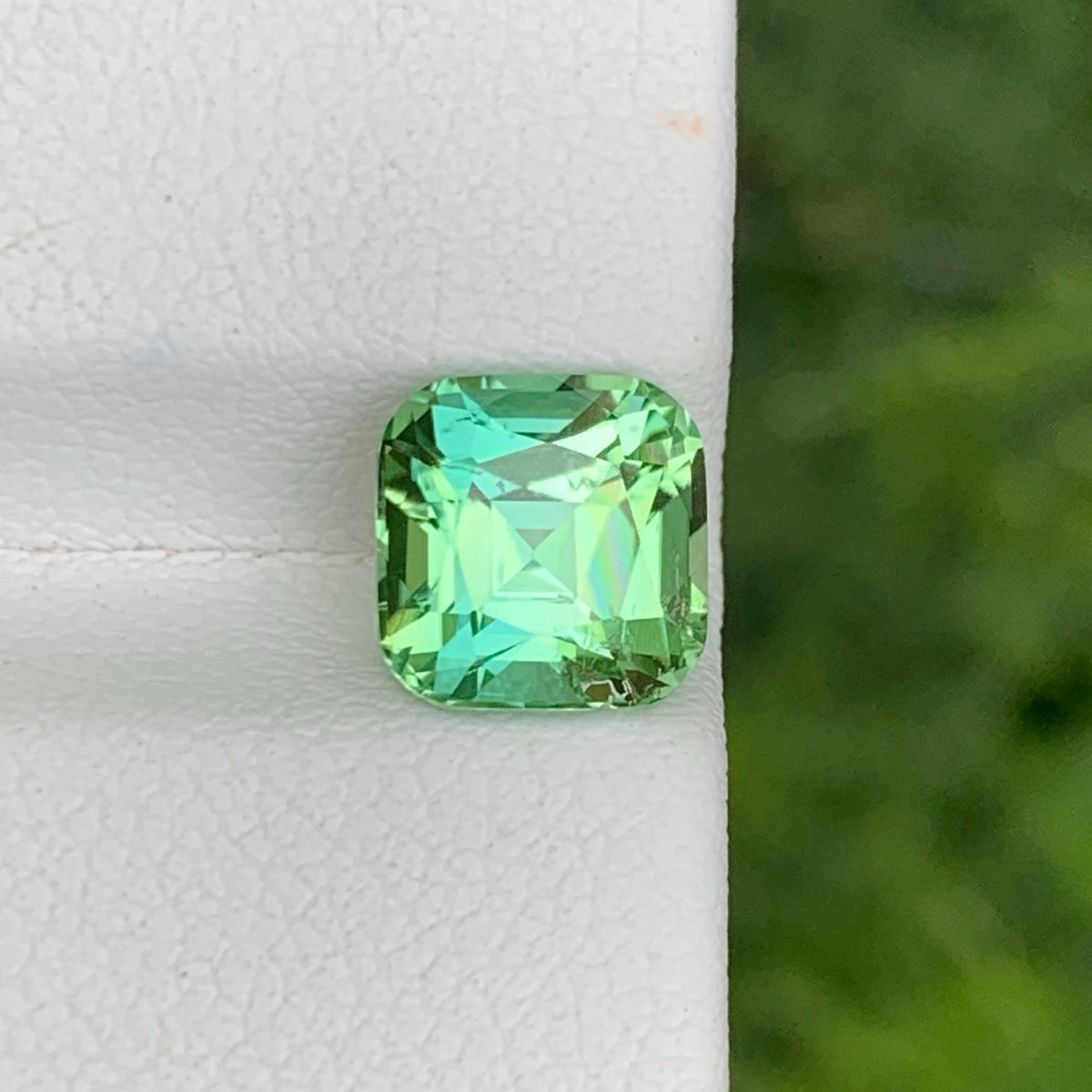 A cushion-cut green gemstone with a vibrant mint and turquoise hue, displayed on a textured white surface with a blurred green background.