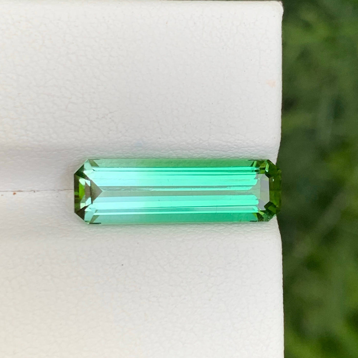 A rectangular emerald-cut green gemstone displayed on a white textured stand, with a blurred green background.