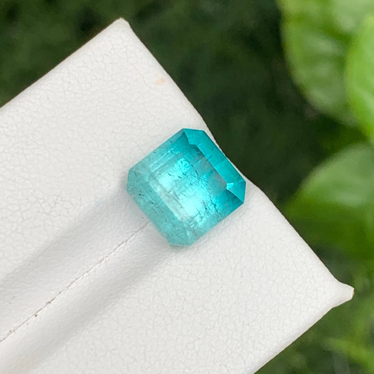 A faceted emerald-cut turquoise gemstone displayed on a white textured jewelry stand, with a soft-focus background of green leaves.