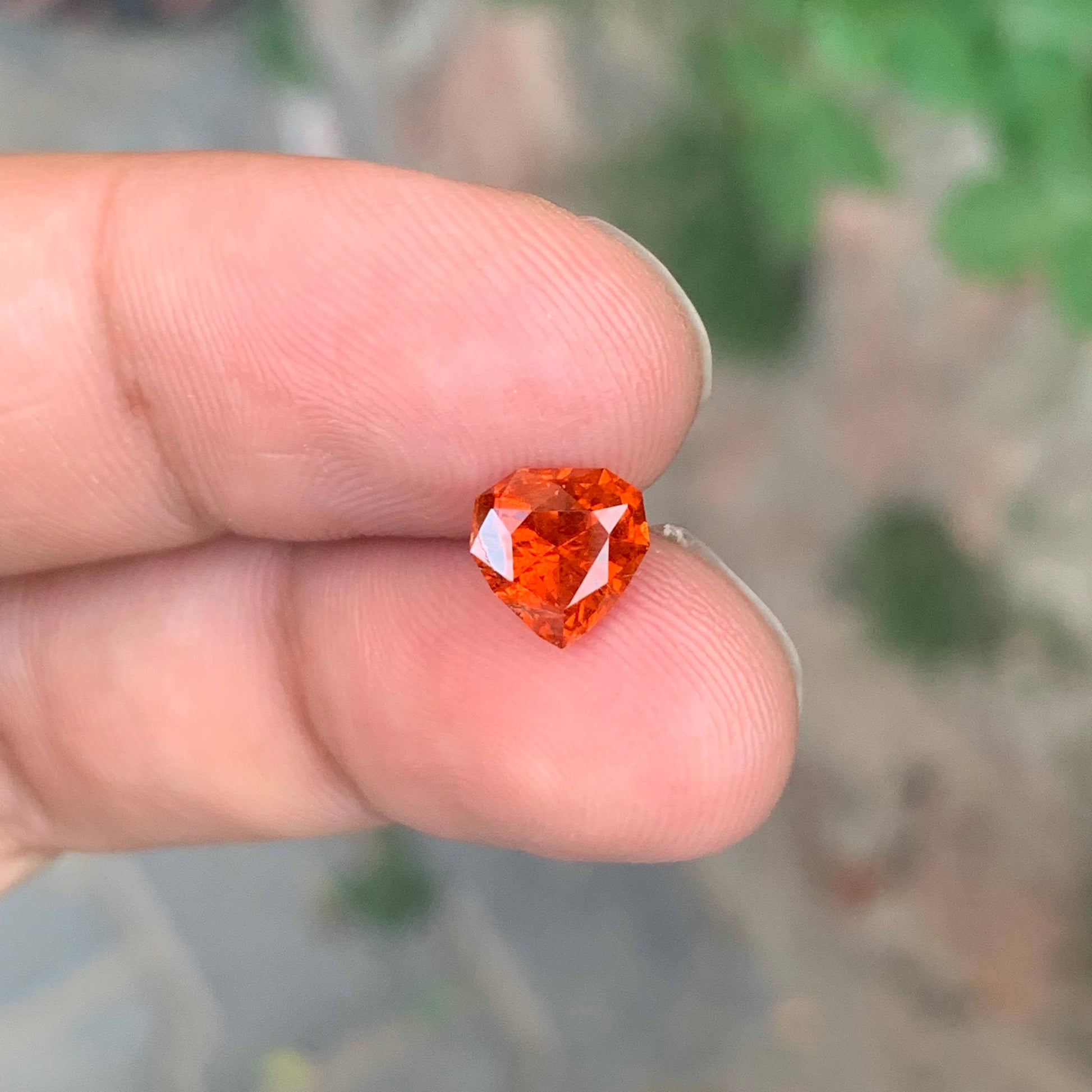 Heart-shaped red gemstone held between fingers with a blurred natural background