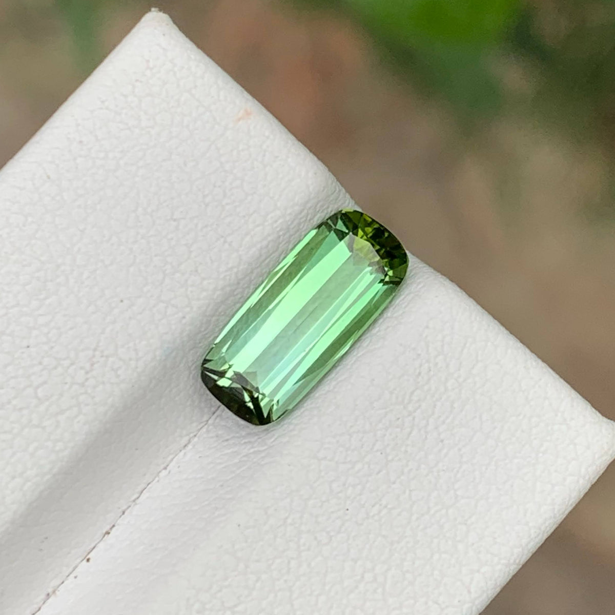 A rectangular emerald-cut green gemstone with faceted edges, displayed on a textured white jewelry stand with a blurred natural background.