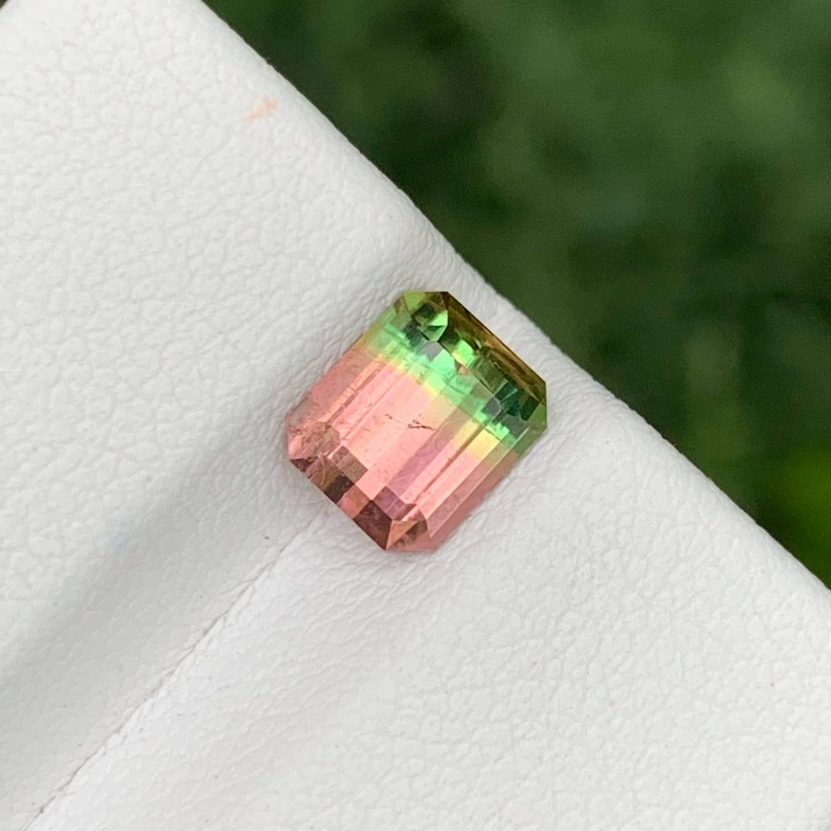 A small rectangular faceted gemstone with a pink-to-green iridescent color shift, resting on a white textured surface with a blurred green background.