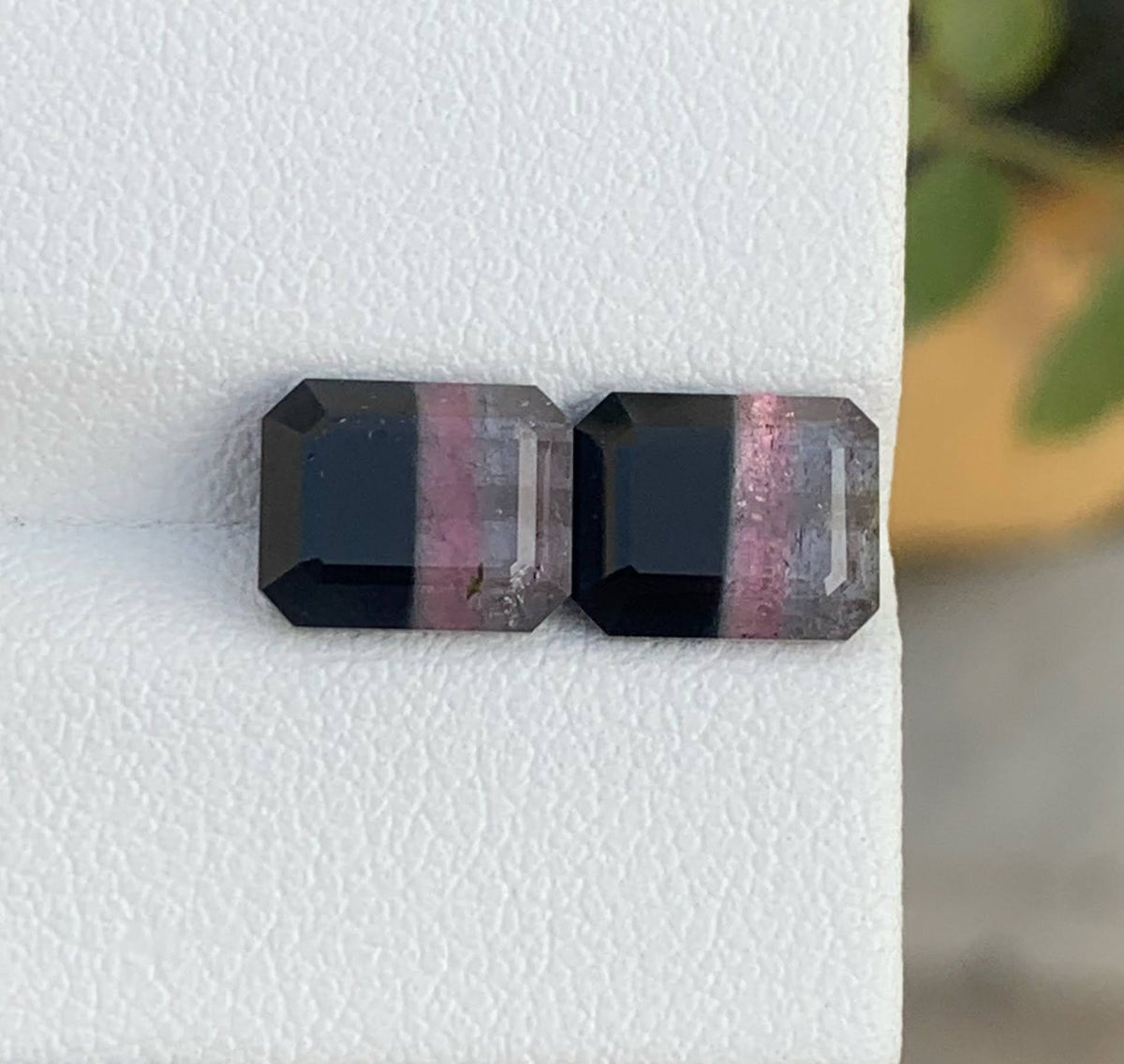 Two small rectangular gemstones with a black body and pinkish-purple banding, displayed on a white textured surface.